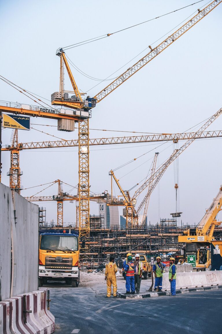 Photograph of a large Middle Eastern construction site at dusk with multiple yellow Potain tower cranes, scaffolding, heavy vehicles, and a group of workers in high-visibility vests gathered in the foreground