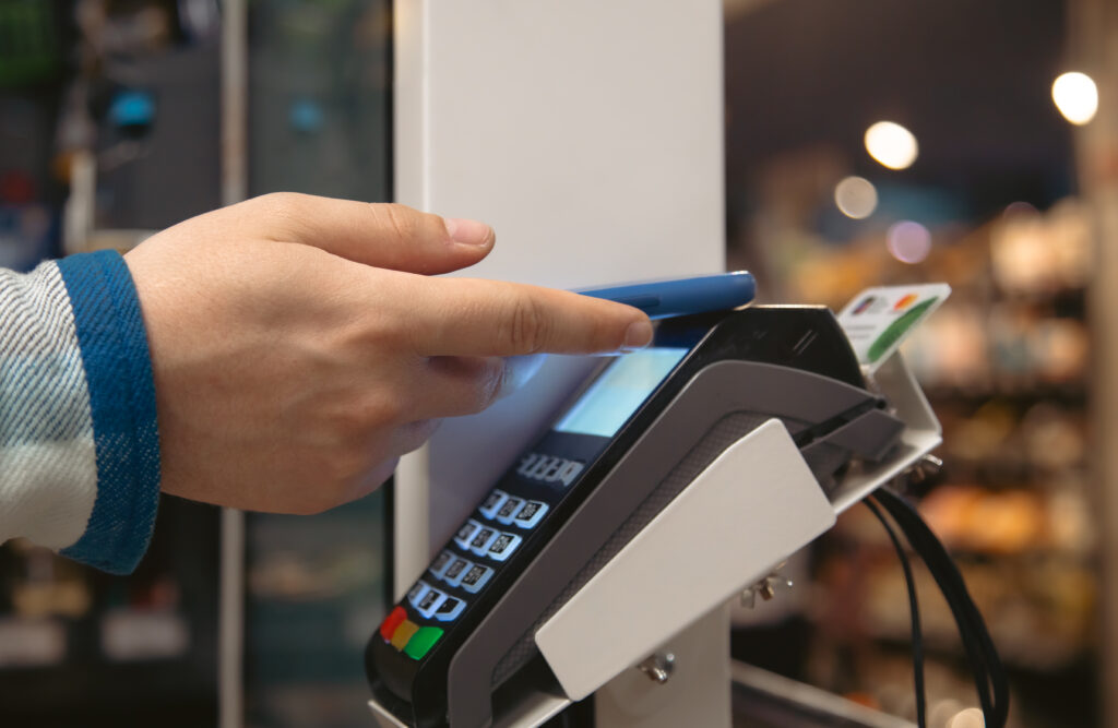 Close-up photograph of a person tapping a smartphone against a POS payment terminal to make a contactless mobile payment in a retail store, with a credit card also visible in the terminal's card slot